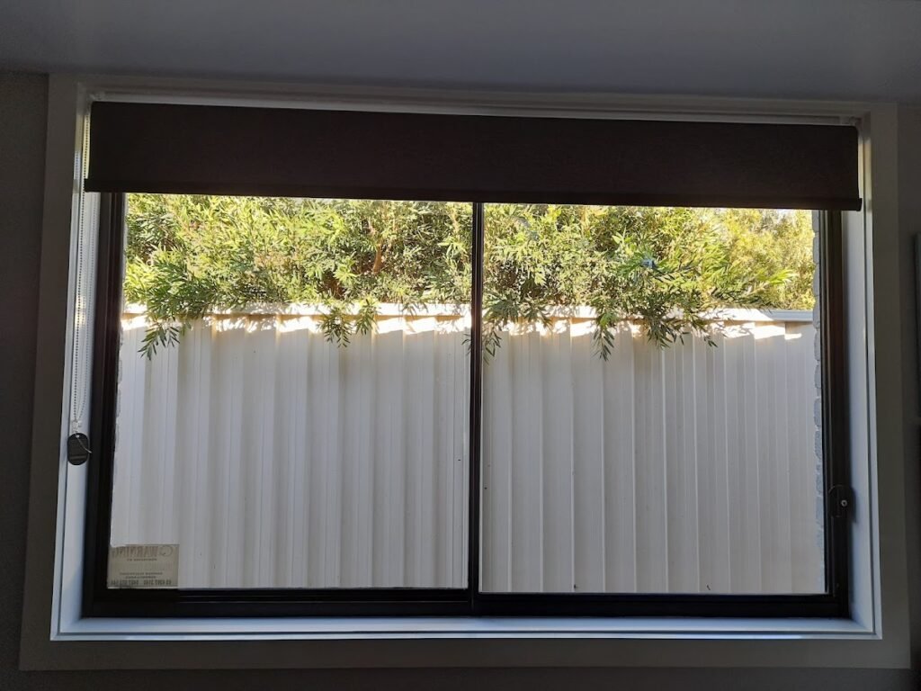 Clean streak-free sliding window with dark roller blind, overlooking a corrugated colorbond fence and greenery